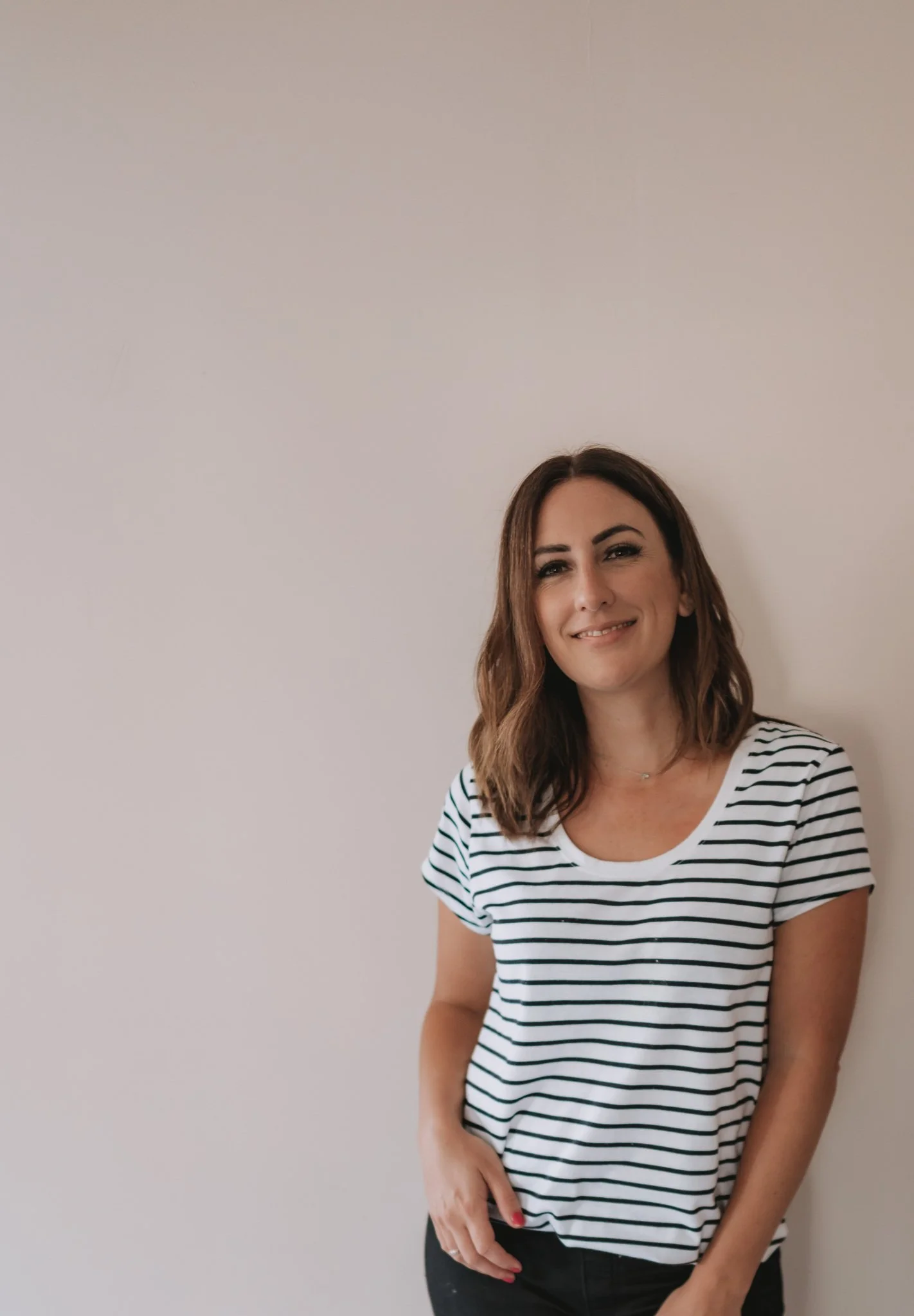 A woman with shoulder-length brown hair, wearing a white and black striped t-shirt, standing against a plain white wall and smiling softly at the camera.