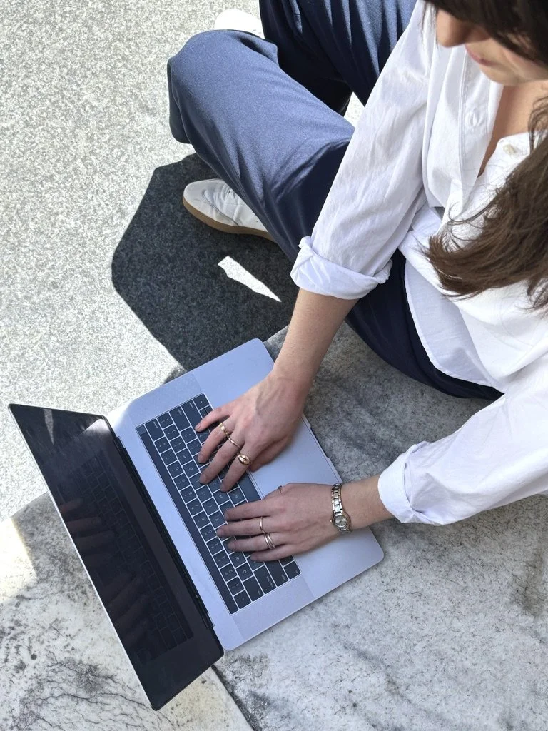 Person sitting on concrete ground using a silver laptop, wearing a white shirt, navy pants, white shoes, and a watch.