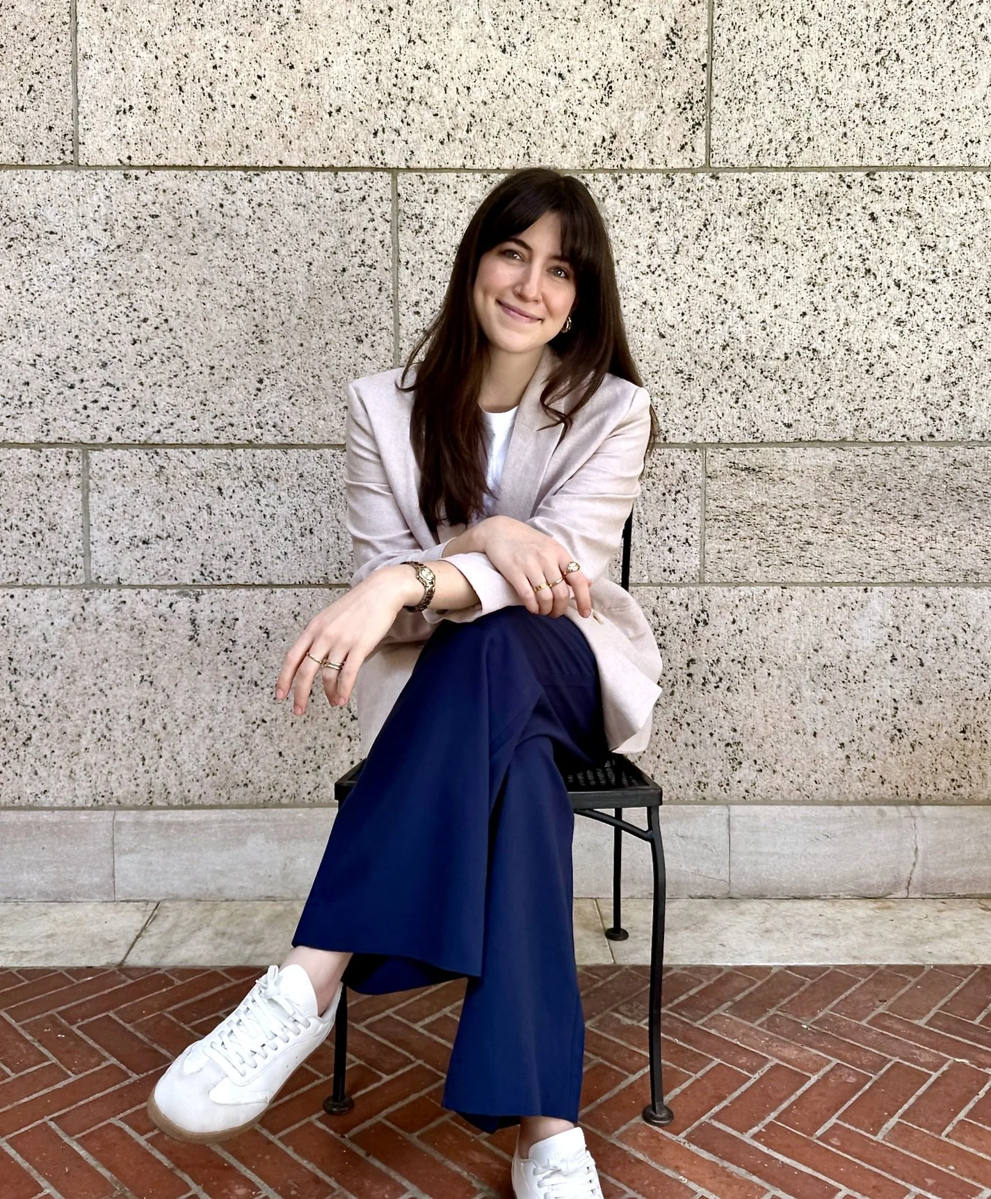 A young woman with long dark hair sitting on a black metal chair against a beige textured stone wall, smiling, wearing a light beige blazer, white shirt, navy blue wide-leg pants, white sneakers, and gold jewelry.