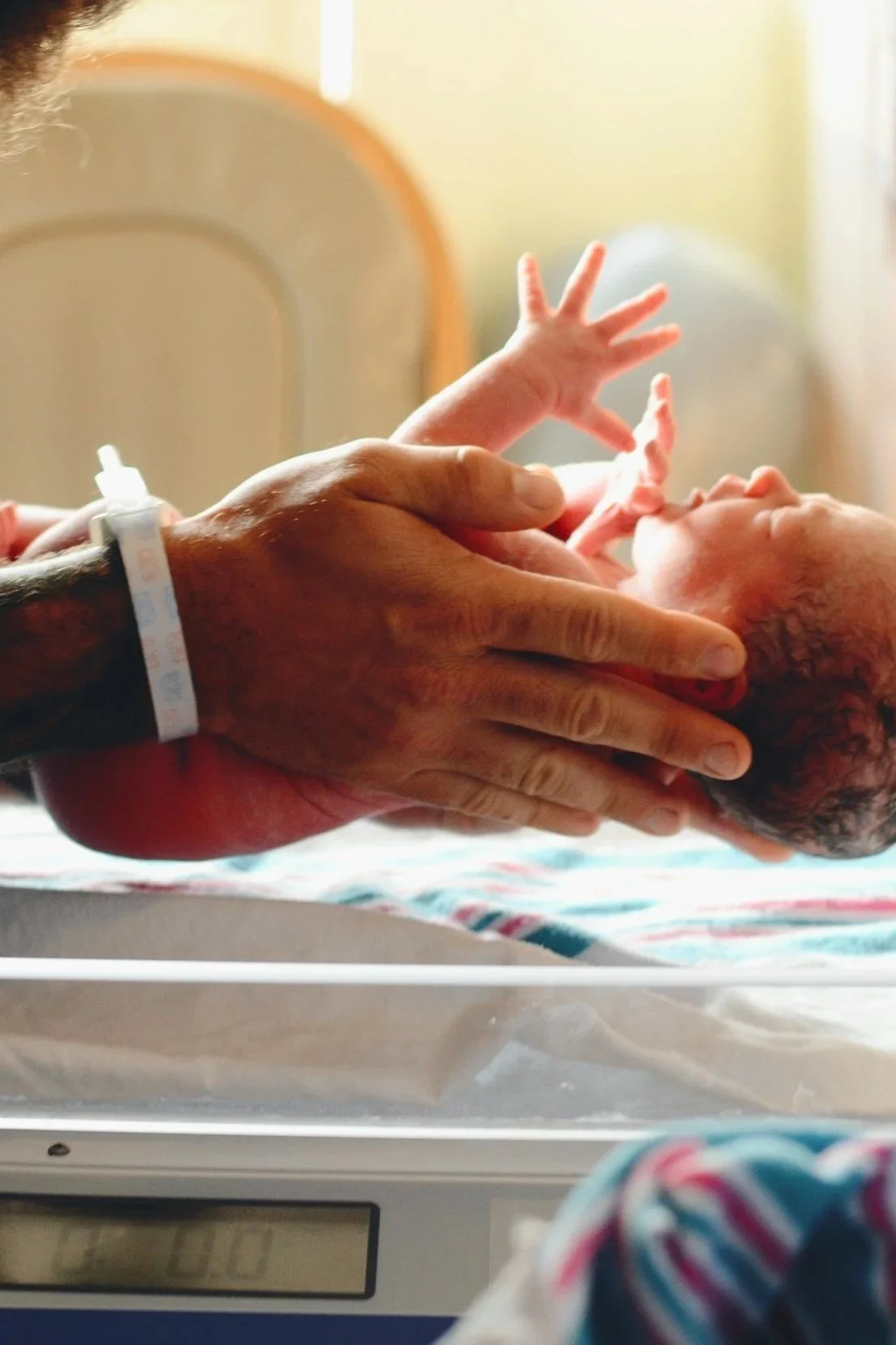 A newborn baby lying on a hospital scale with an adult holding the baby's head and body. The adult has a hospital ID wristband and an IV in their arm. The background is softly lit and out of focus.