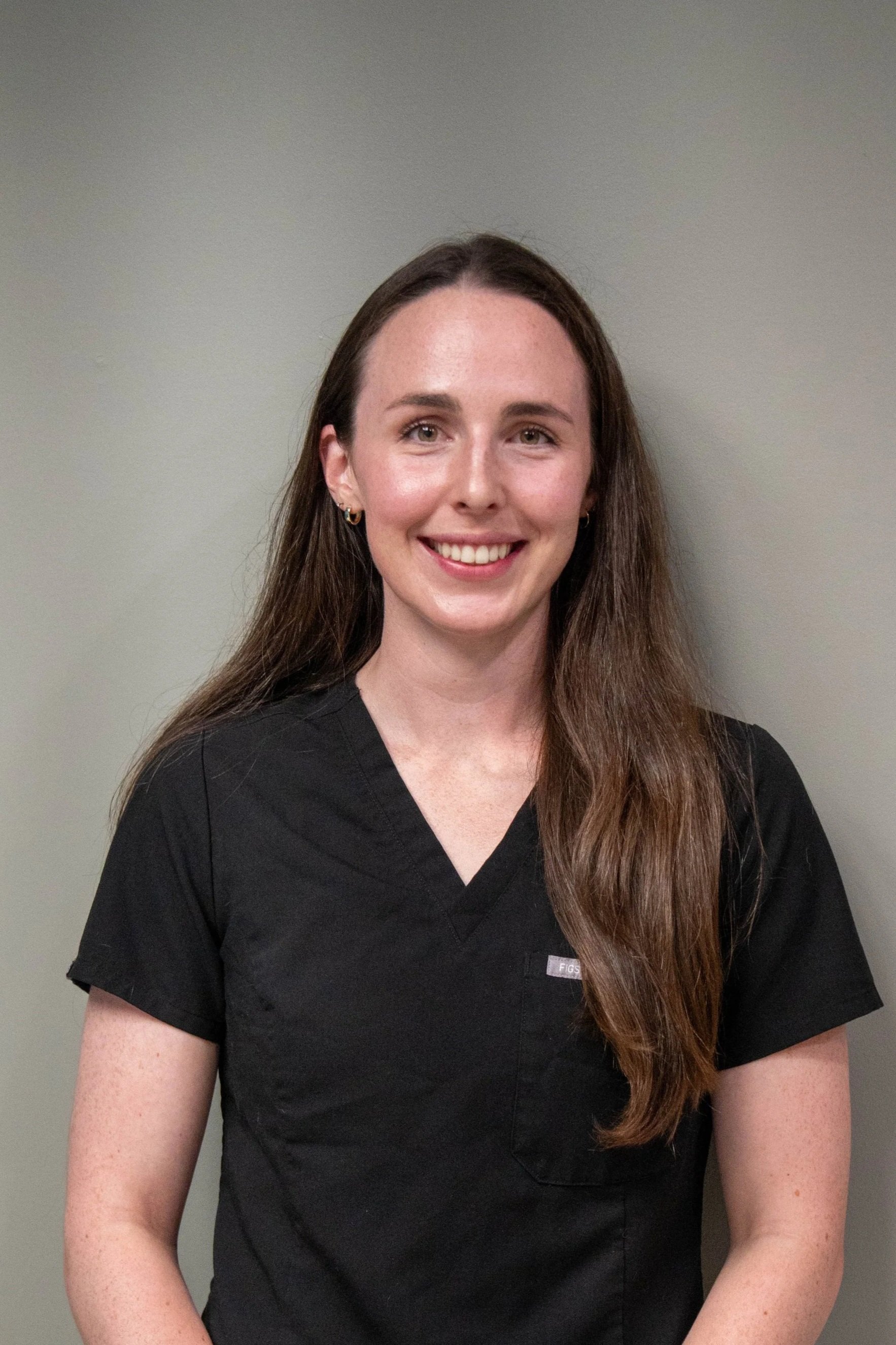 A smiling woman with long brown hair wearing a black medical scrub top standing against a light gray wall.