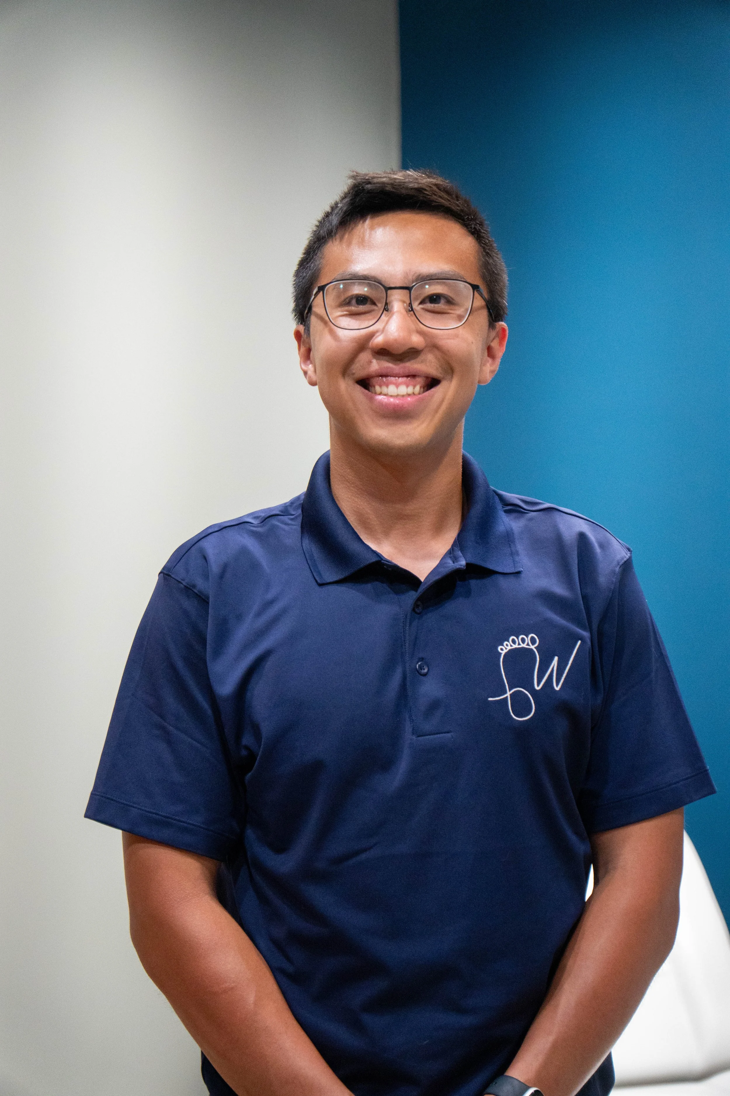 A smiling man wearing glasses and a navy blue polo shirt with a logo on the left chest, standing indoors against a background with white and blue walls.