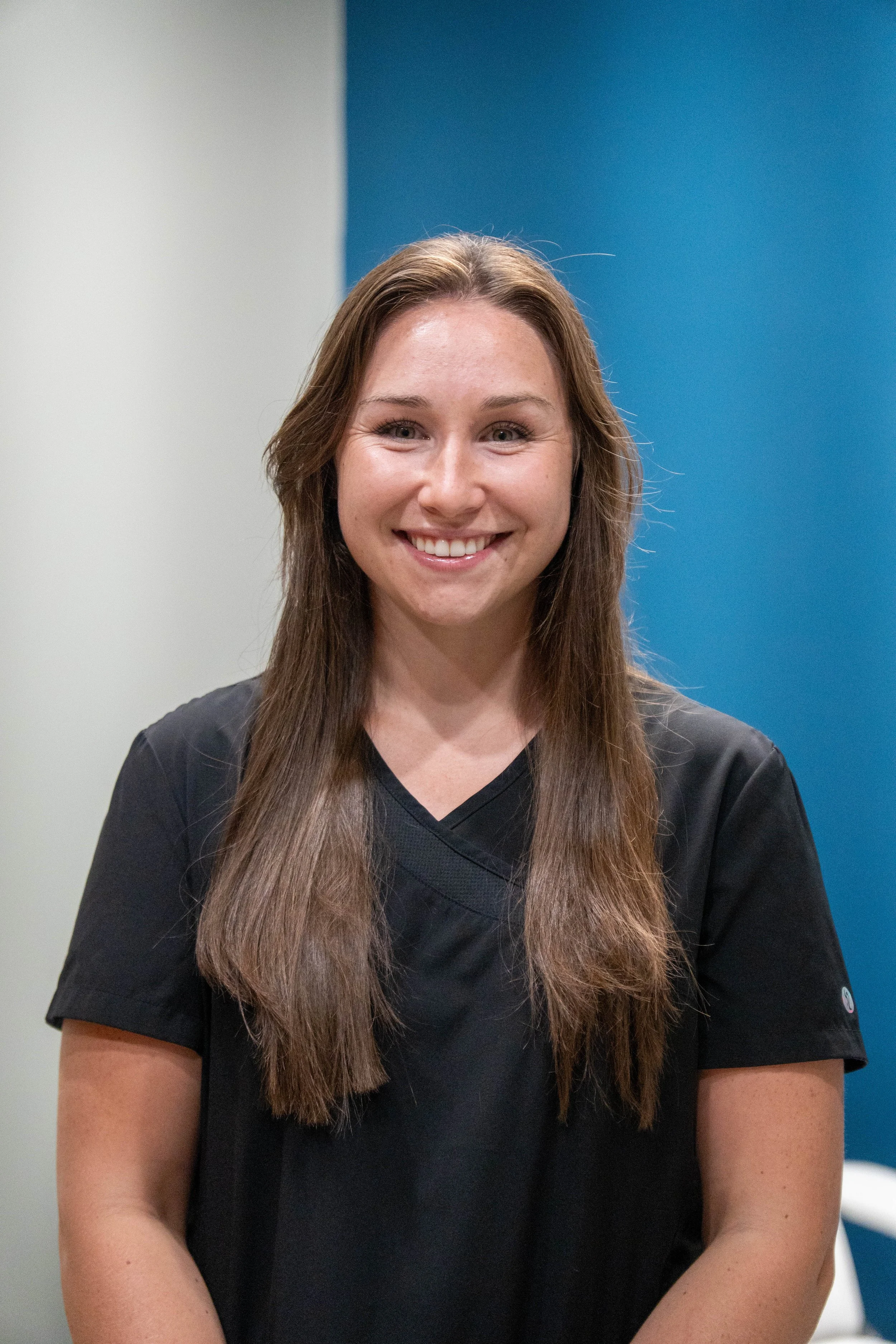 A young woman with long brown hair, smiling, wearing a black medical scrub top, standing against a background with a white wall on the left and a blue wall on the right.