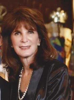 A woman with shoulder-length brown hair, wearing a black shiny jacket and a pearl necklace, smiling in an indoor setting with warm lighting.