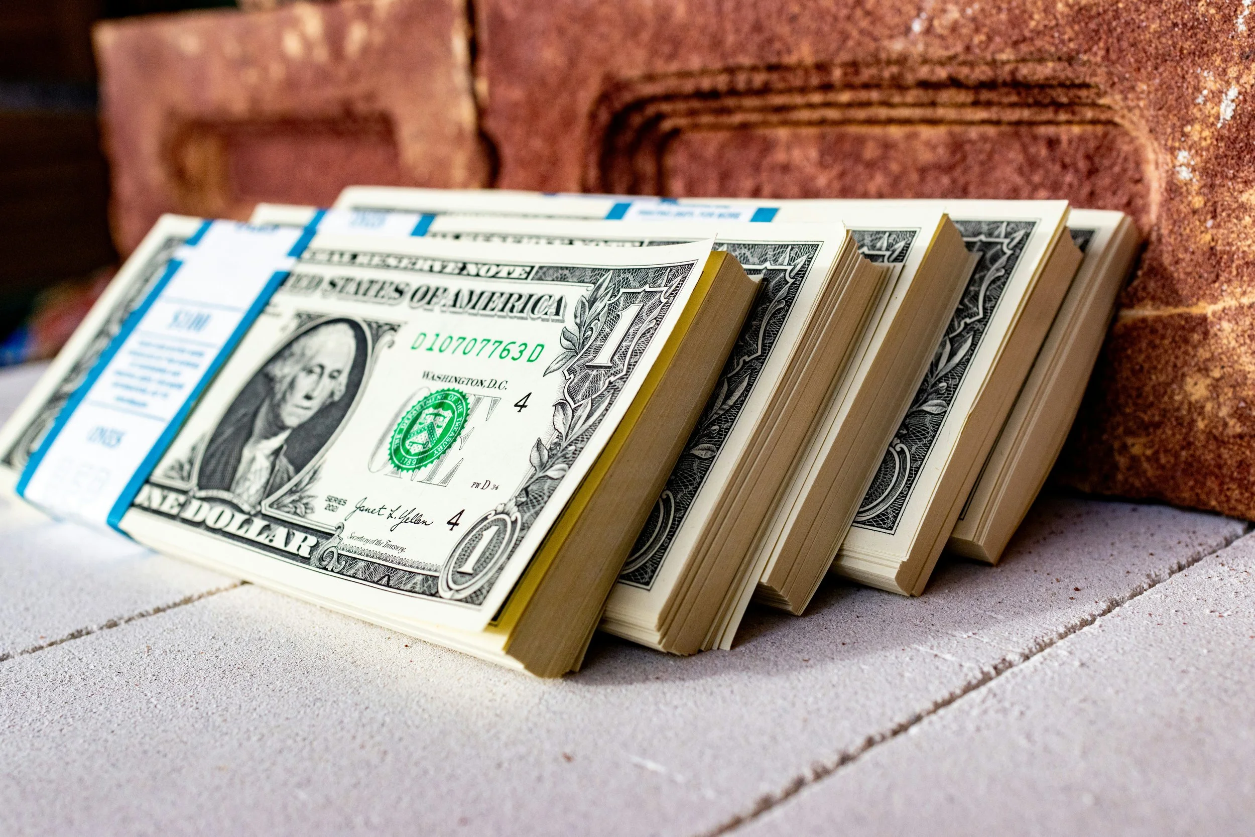 Stacks of US one-dollar bills bundled with blue bands, positioned on a surface, with a brick wall in the background.