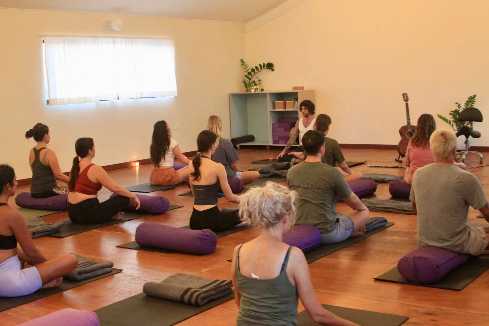 People participating in a yoga or meditation class in a bright studio, sitting cross-legged on mats with bolsters. Practicing Balance, Therapy, Integrated Guidance, Coaching, Yoga, Workshop Facilitation and Art and Music Therapy.