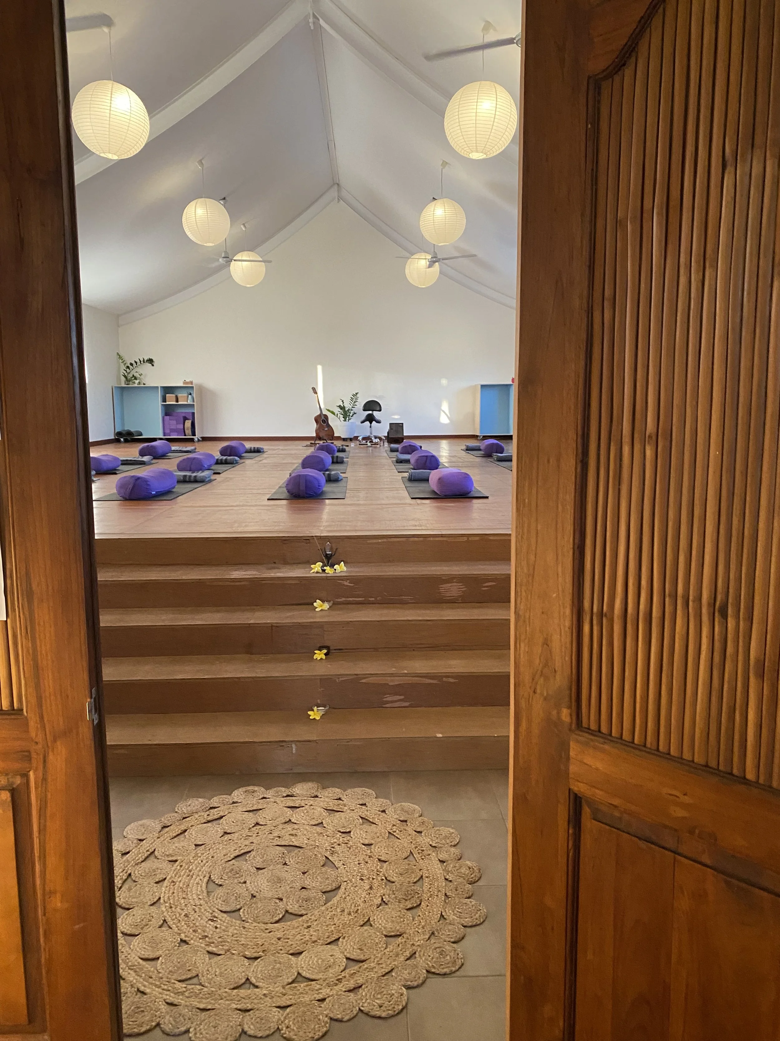 View through open wooden door into a minimalist yoga studio with wooden floor, purple cushions on yoga mats, hanging paper lanterns, and a guitar on the floor. Ready for Music therapy, Balance, Therapy, Integrated Guidance, Coaching, Yoga, Workshop