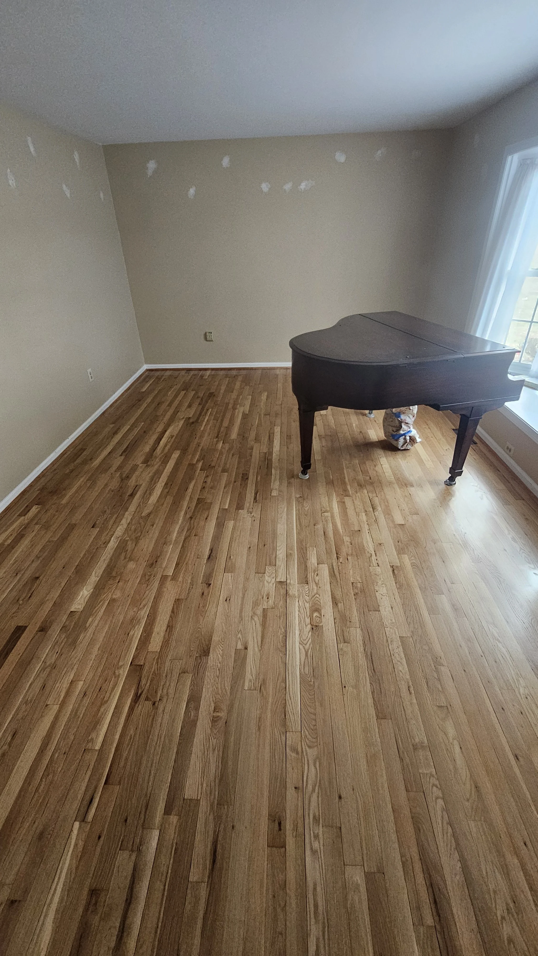 Empty room with hardwood flooring, beige walls with some white paint patches, a window with curtains, and a black grand piano.
