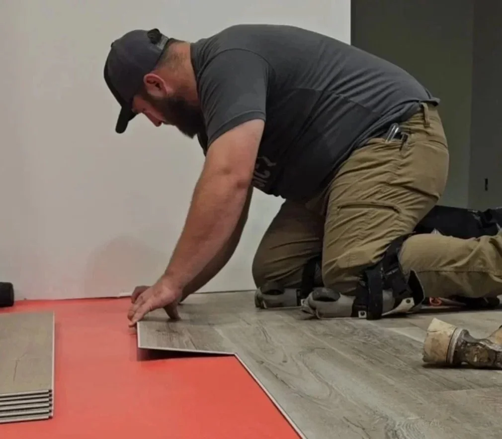 A man kneeling on the floor, installing a laminate or vinyl flooring next to a section of red underlayment.