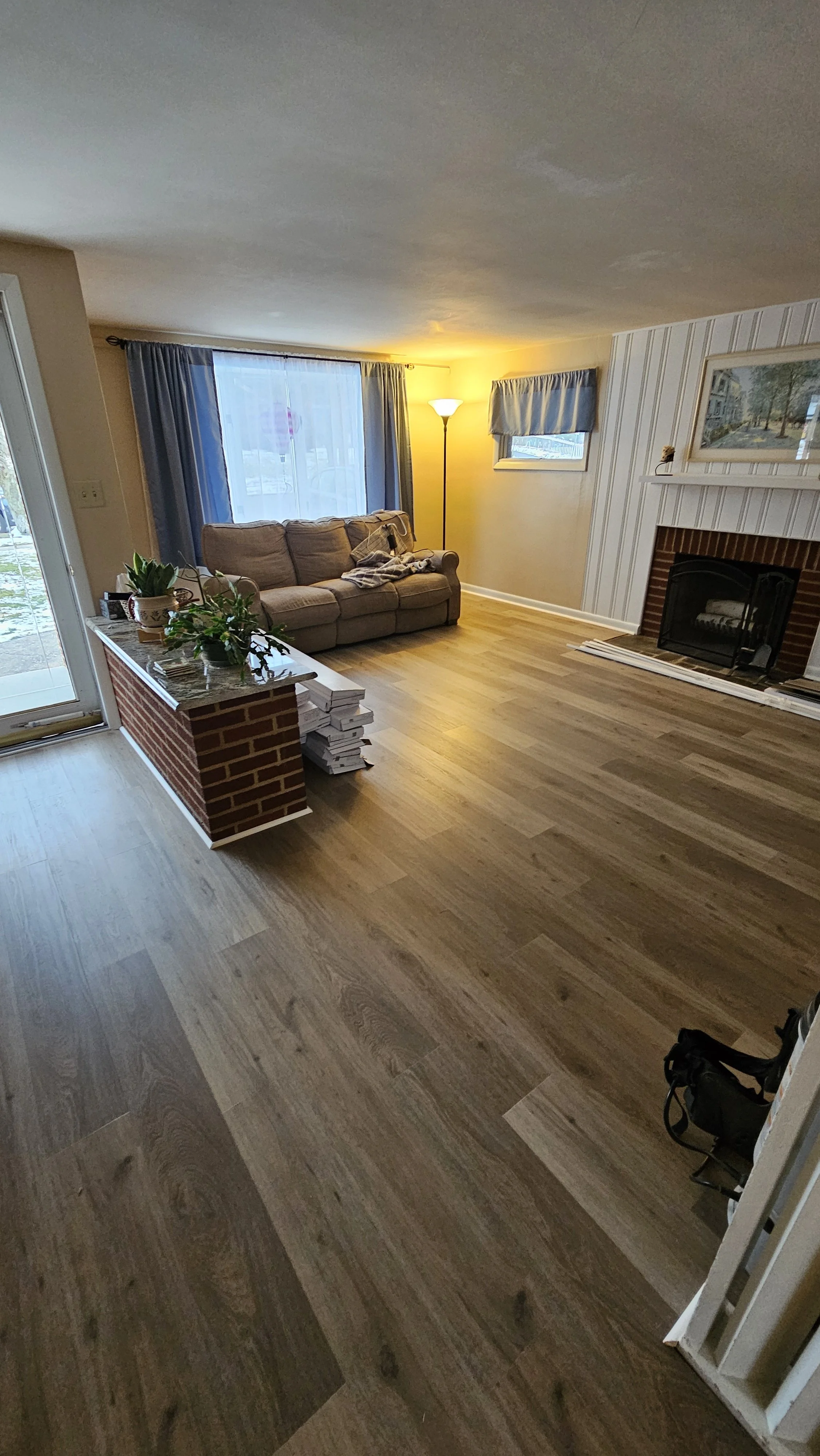 Living room with a beige couch, a brick fireplace, a floor lamp, and windows with curtains. There is a small table with a plant near the door, and a backpack on the floor.