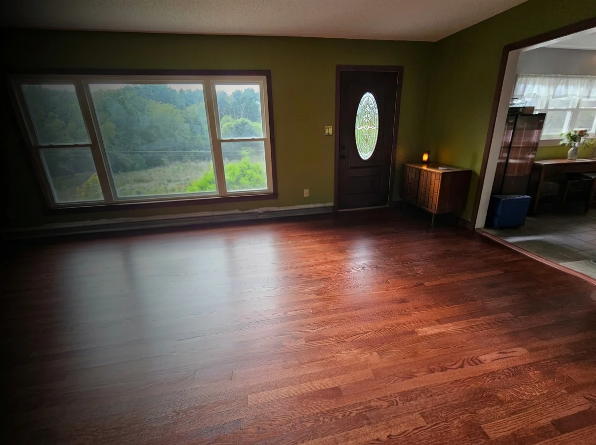 Empty living room with large window overlooking trees, dark wood floor, green walls, front door with oval glass insert, small wooden side table with lit candle, and an open doorway leading to a dining area with a table, chairs, and a window with curtains.