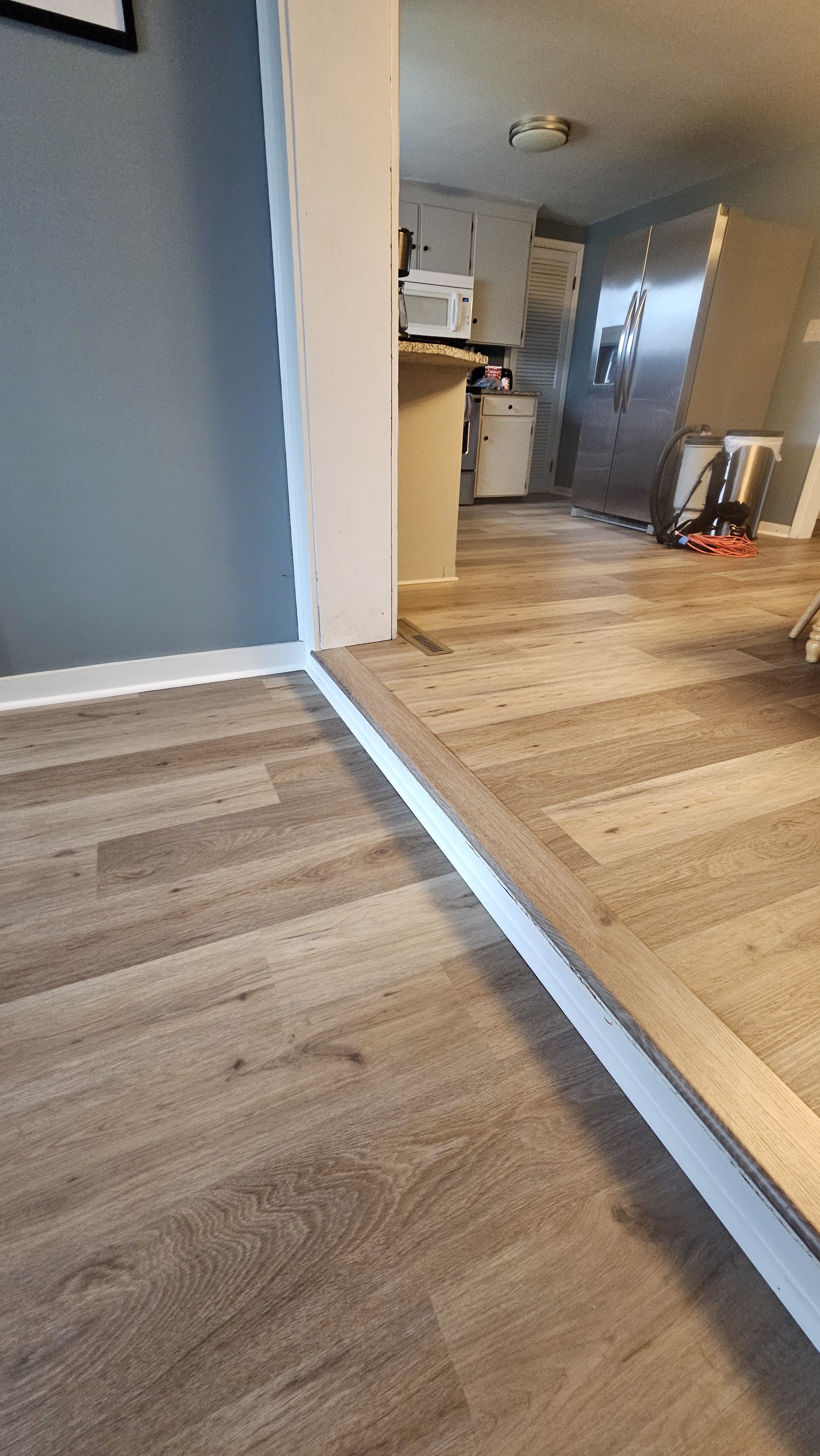 A photo of a kitchen and living area with wood flooring, showing a transition strip between the two types of flooring, with appliances including a microwave, refrigerator, and vacuum cleaner visible in the background.