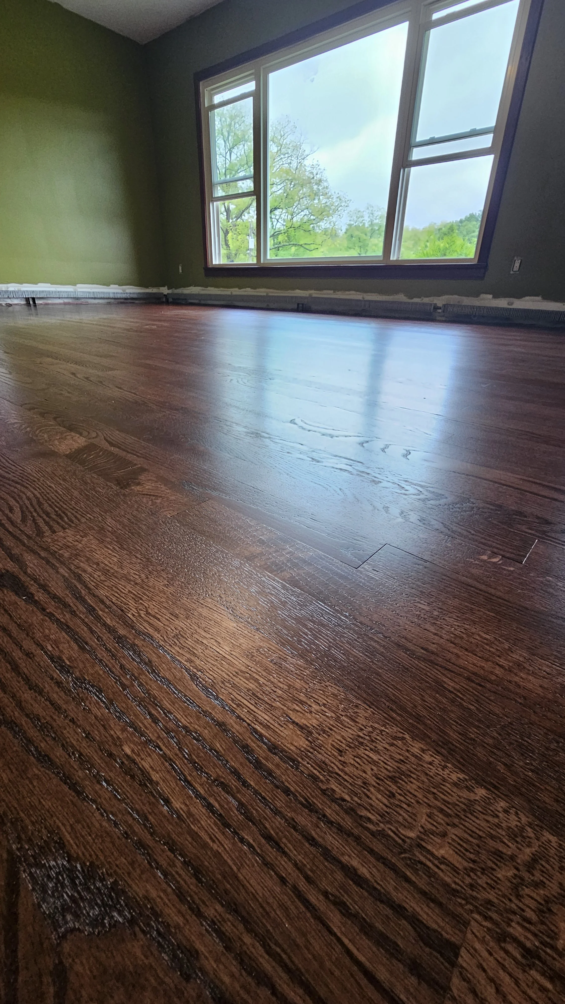 Photo of a clean, shiny hardwood floor in a room with green walls and a large window showing trees outside.