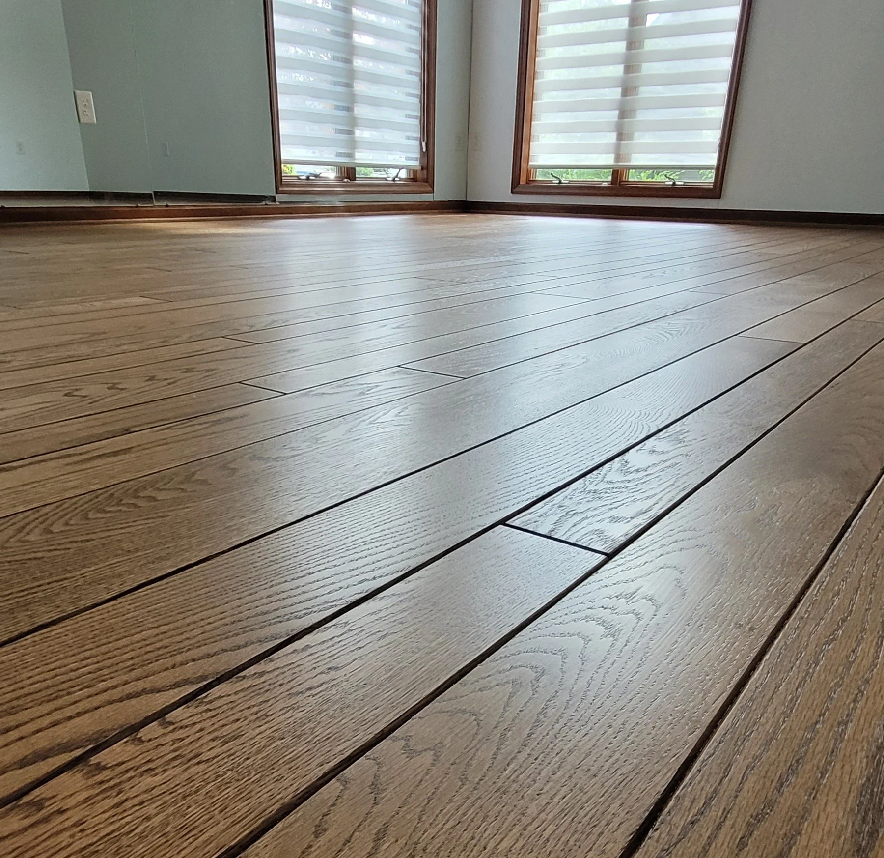 Close-up of a wooden floor with wide planks and visible wood grain, near a wall with a white electrical outlet and two large windows with wooden frames and striped blinds.