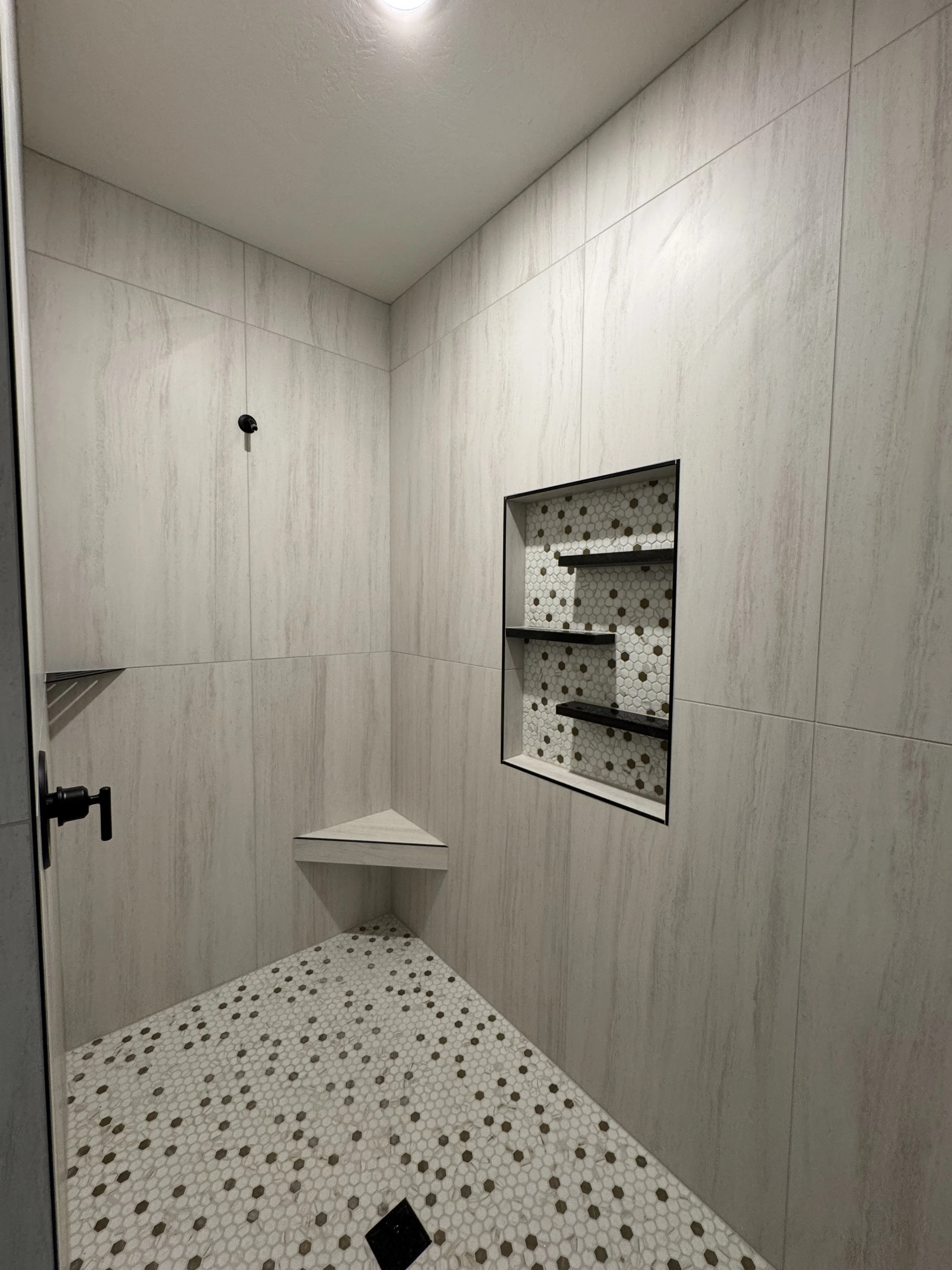 Empty shower with beige tiled walls, built-in shelves with a honeycomb tile background, and a corner seat.