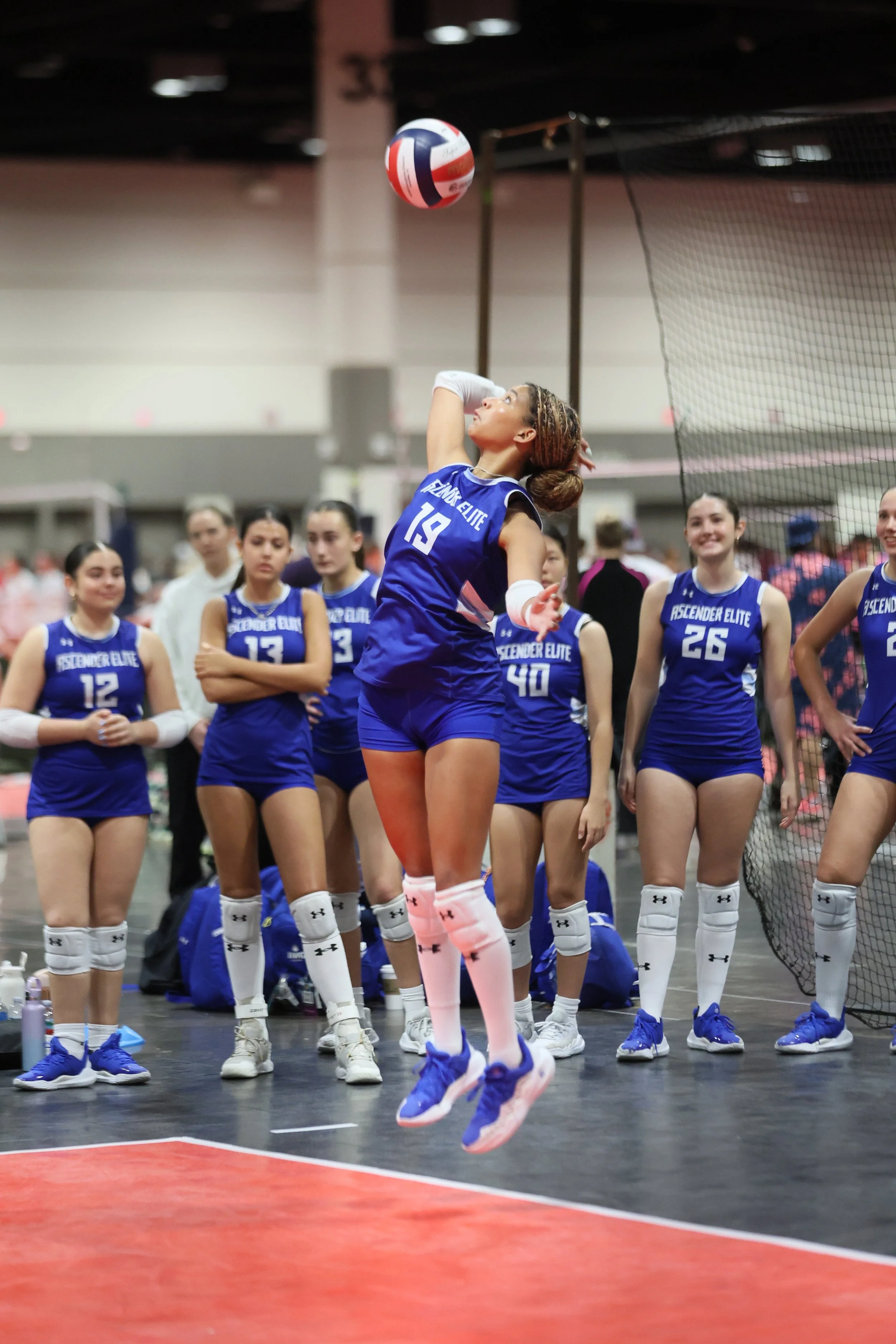 A women's volleyball team wearing blue uniforms with 'Ascender Elite' written on them, gathered together, with one player in mid-air preparing to serve, inside an indoor gymnasium.