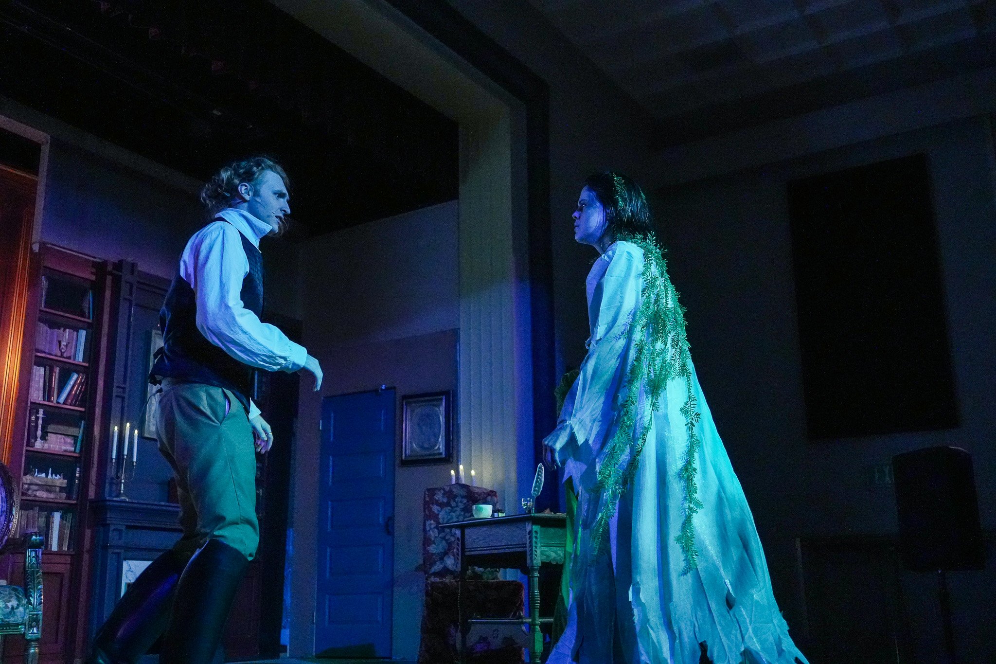 A scene from a theatrical production showing a man and a woman facing each other in a dimly lit room. The man is dressed in historical attire with high boots and a vest, while the woman wears a flowing white dress with green foliage accents. The room