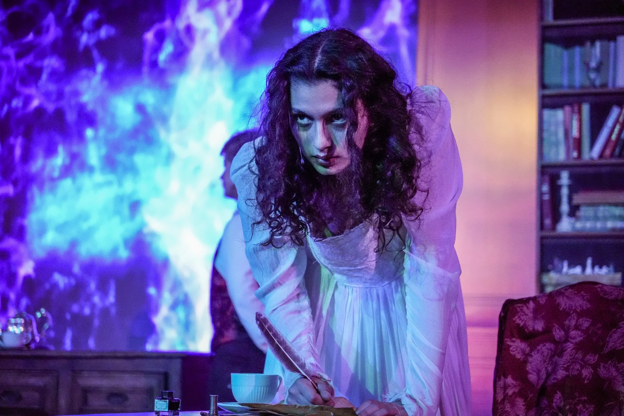 A woman with long, curly hair and dramatic makeup leans over a table, holding a quill pen, in a room with purple and blue lighting and bookshelves in the background.