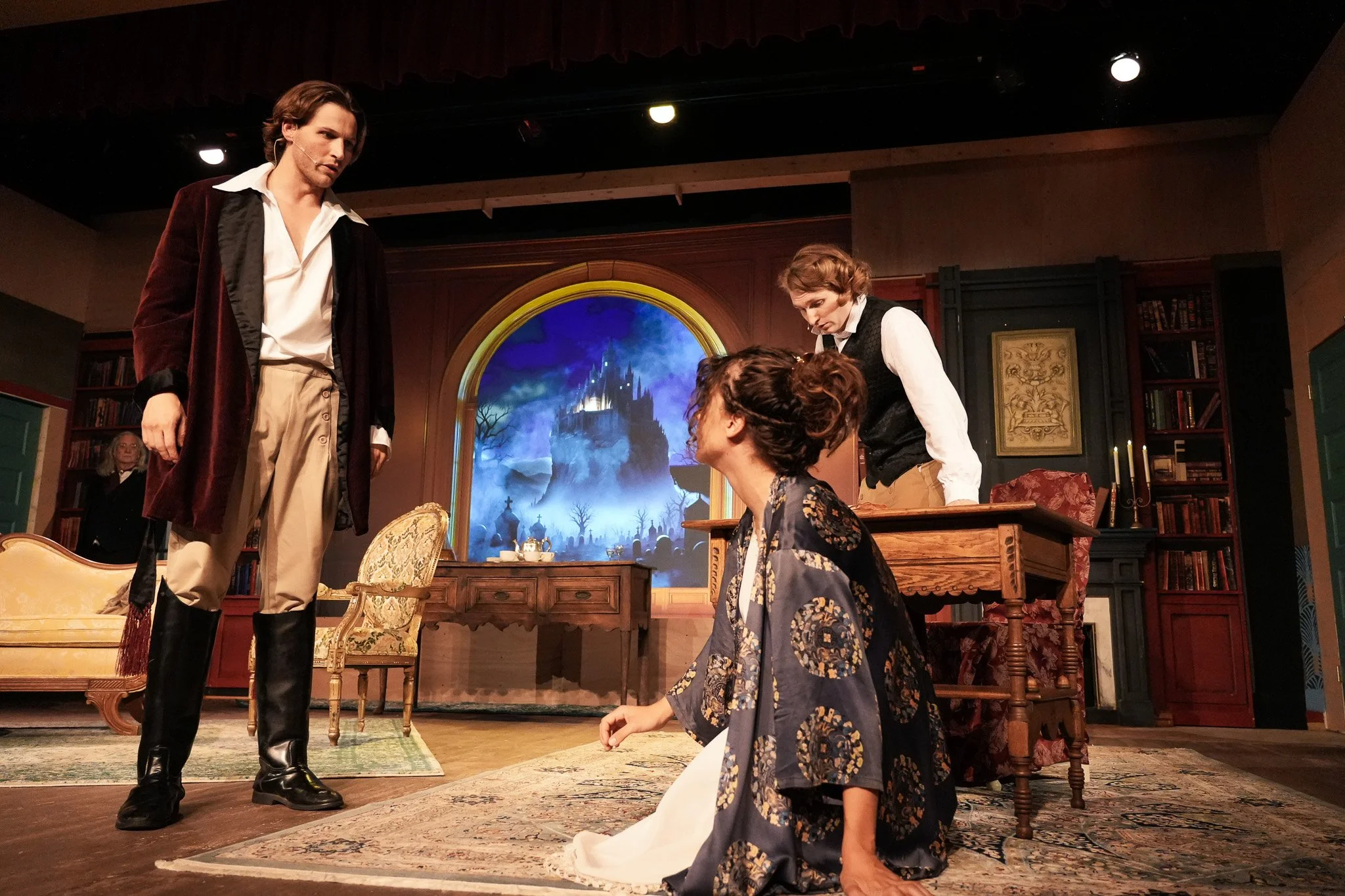 Four actors on stage in a theatrical play, with a backdrop resembling a castle on a moonlit night, and a room set with old-fashioned furniture.