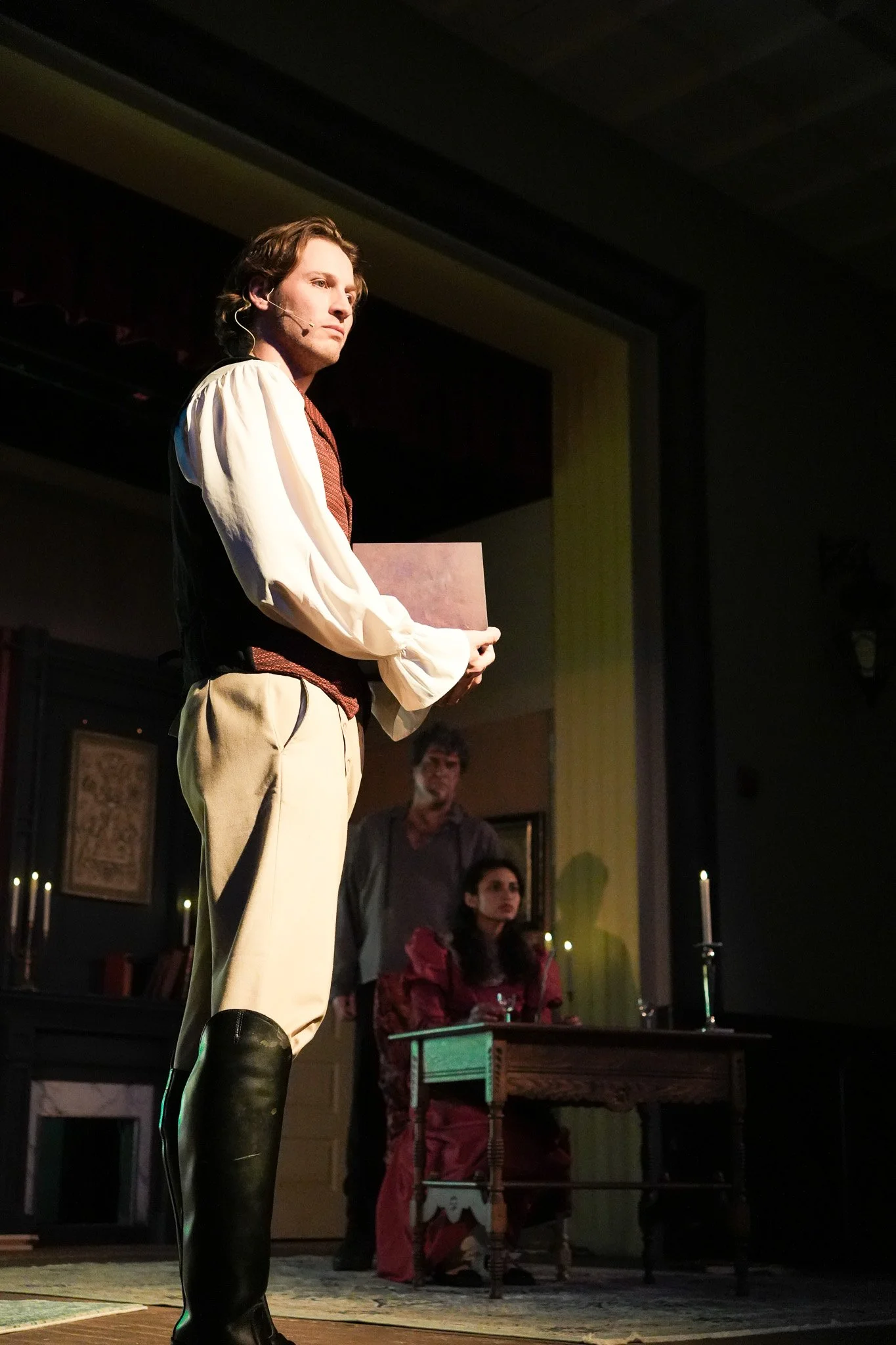 A young man dressed in historical costume stands on stage, holding a document, with three other people seated and standing behind him in a theatrical scene.