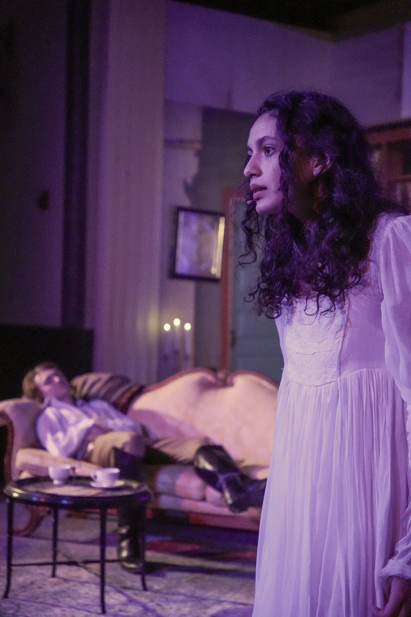 A woman with curly hair dressed in a light pink nightgown stands in a dimly lit room with purple lighting, while a person with light brown hair and pale skin lays on a vintage sofa in the background.