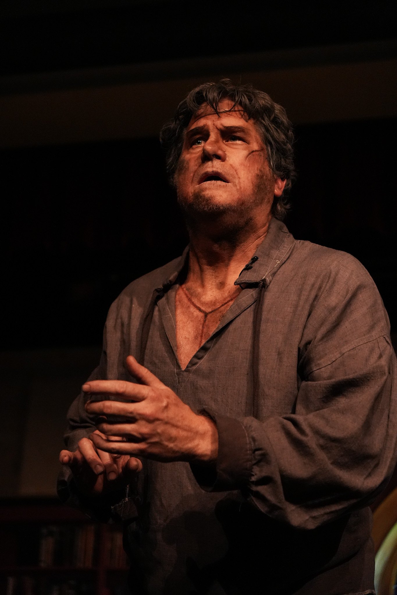 A man with gray hair and a beard, wearing a brown shirt, appears distressed, with his hand on his chest, in a dimly lit setting.