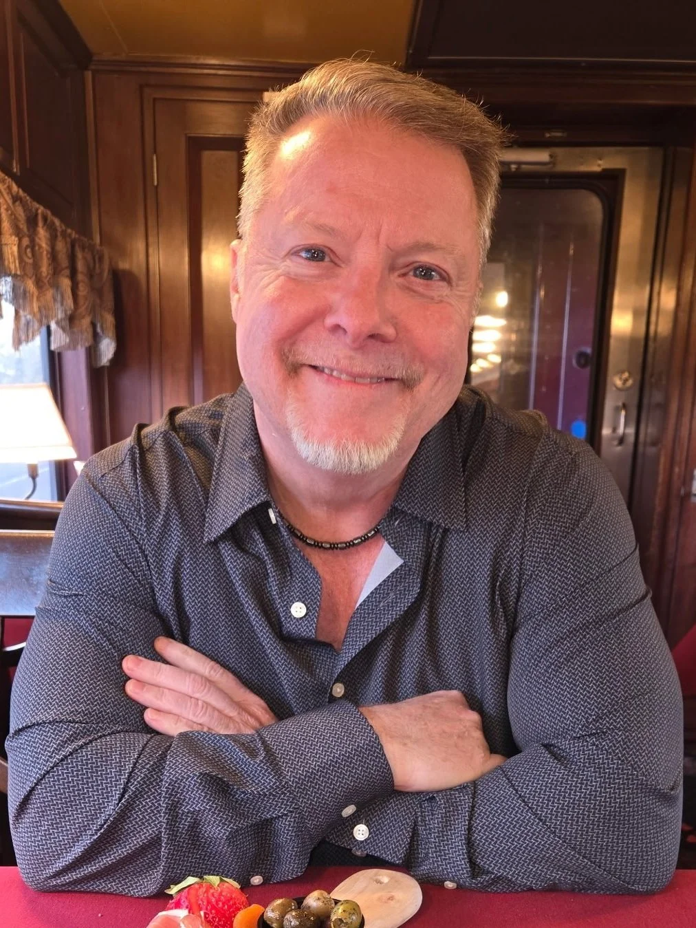 A smiling man with gray hair and a beard sitting at a table with a plate of strawberries, olives, and small vegetables in front of him in a wooden-paneled room.