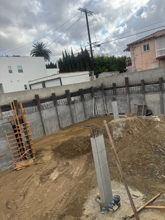 Construction site with rebar and concrete pillars being built, surrounded by dirt and a concrete wall with houses and trees in the background under a cloudy sky.