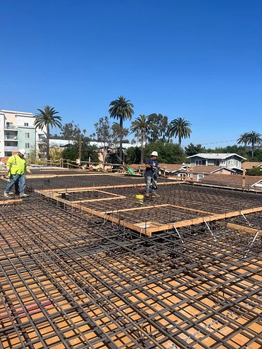 Construction workers laying rebar on a building foundation, with wooden frames, under a clear blue sky surrounded by palm trees and nearby houses.