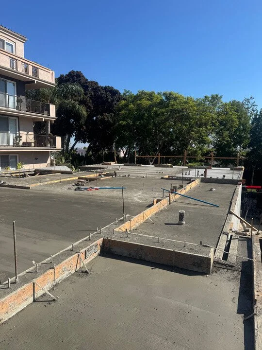 Construction site with freshly poured concrete on a building's upper floor, with construction tools and safety barriers, next to an apartment building and green trees under a clear blue sky.
