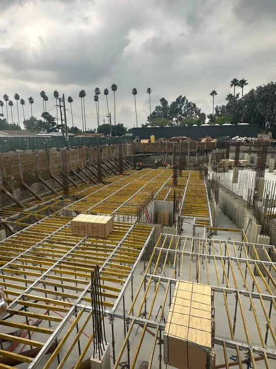 Construction site with wooden and metal framework, under cloudy sky, with palm trees in the background.