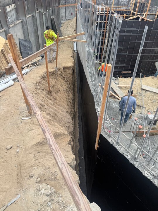 Construction site with workers installing waterproofing on a vertical concrete wall, scaffolding, and safety barriers.