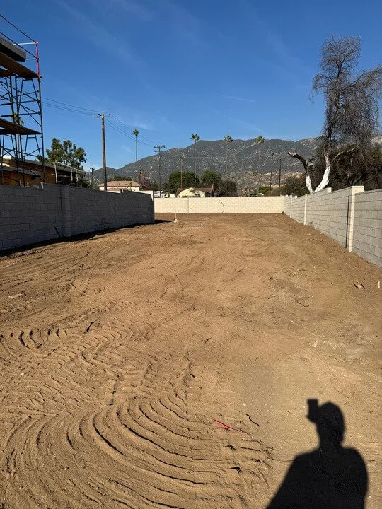 A dirt lot enclosed by gray concrete walls with a mountain range and clear blue sky in the background. There is a construction scaffold on the left, a tall dead tree on the right, and a person's shadow in the foreground.