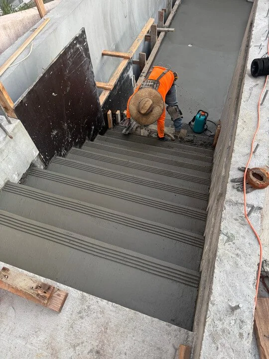 A construction worker is applying concrete to a staircase at a construction site, with wooden formwork and tools around.
