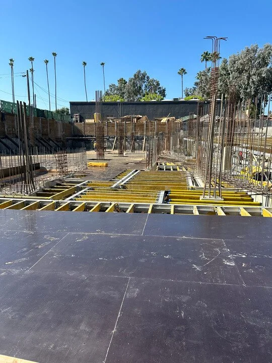Construction site with wood and metal framing for a building, under clear blue skies, with palm trees in the background.