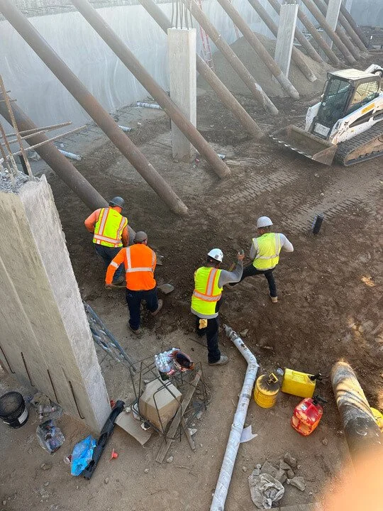 Construction workers in safety vests and helmets working on a dirt area at a construction site with heavy machinery and building structures around.