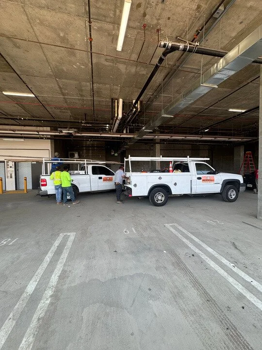 Construction workers inspecting utility trucks in an underground parking garage with exposed ceiling pipes.
