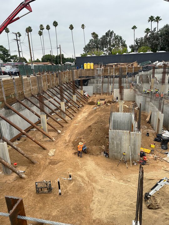 Construction site with workers, concrete walls, and support beams for building foundations, with palm trees and cloudy sky in the background.