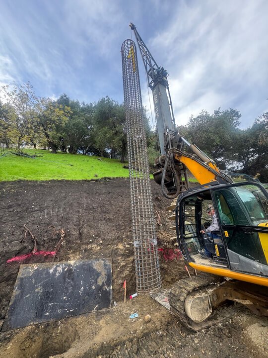 Construction site with a large excavator installing a tall, cylindrical metal reinforcement structure into the ground, with trees and a grassy hill in the background.