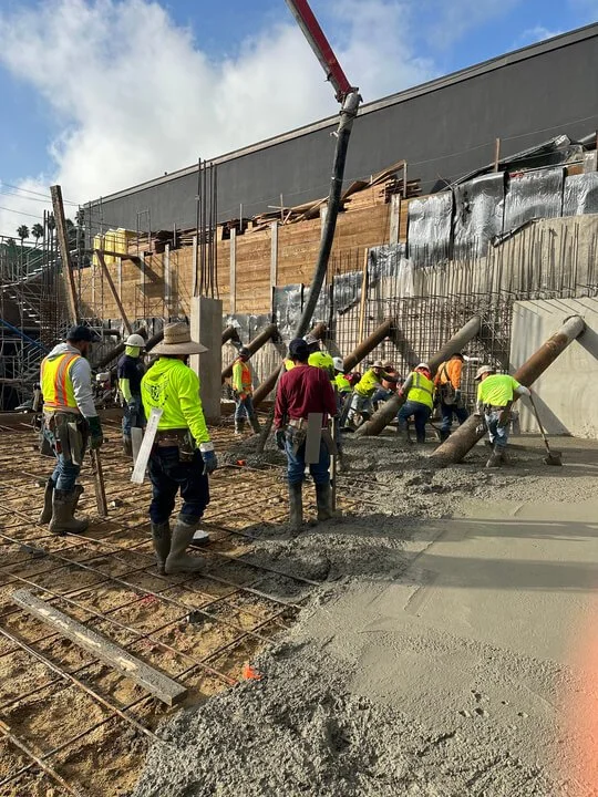 Construction workers in high-visibility vests and hats pouring and leveling concrete for a sidewalk or foundation at a building site.
