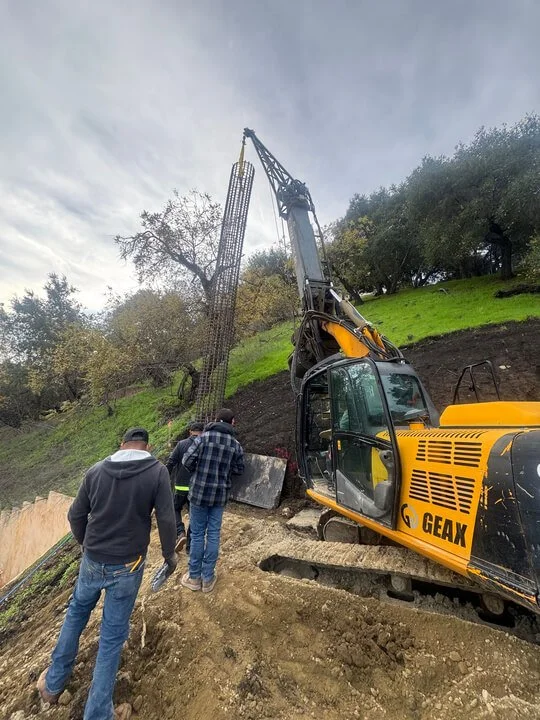 A yellow GEAX excavator is demolishing a hillside with a crane arm extended and a large metal demolition tool. Several people are standing nearby observing the work on a dirt slope with green grass and trees in the background.