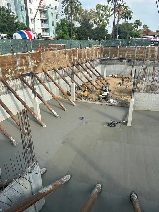 Construction workers pouring concrete on a large foundation with wooden supports and rebar.