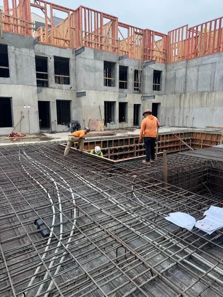 Construction site with steel rebar framework for a building's concrete slab, three workers wearing safety gear working on the rebar, and a partially built multi-story structure in the background.