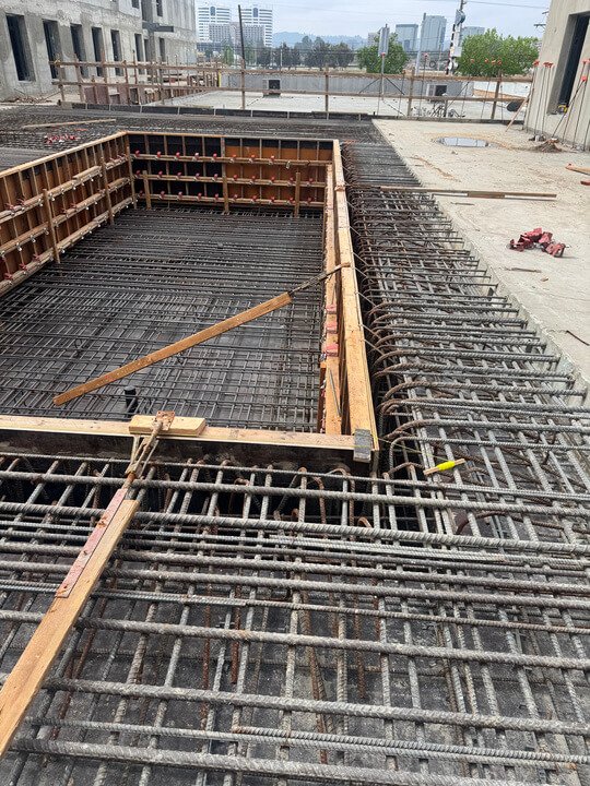 Construction site with rebar framework for a building's foundation, including wooden formwork and metal reinforcement bars.