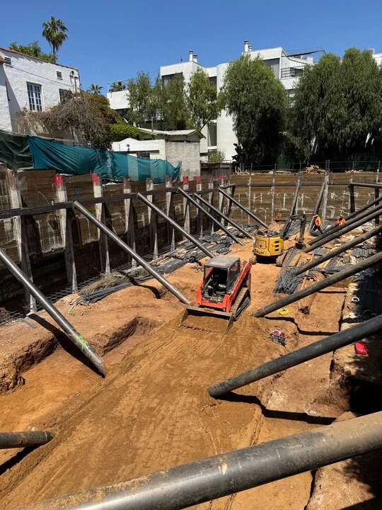 Construction site with dirt and scaffolding, yellow and red compact roller machines, surrounded by trees and residential buildings.