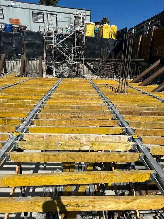 Construction site with yellow wooden formwork and metal supports for pouring concrete, with scaffolding and a building with yellow bins in the background.