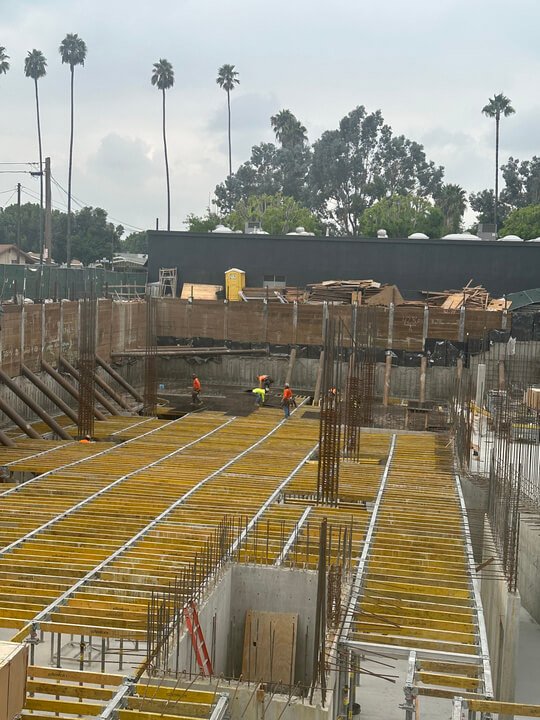 Construction workers on a building site with rebar and wooden formwork, with palm trees and a cloudy sky in the background.