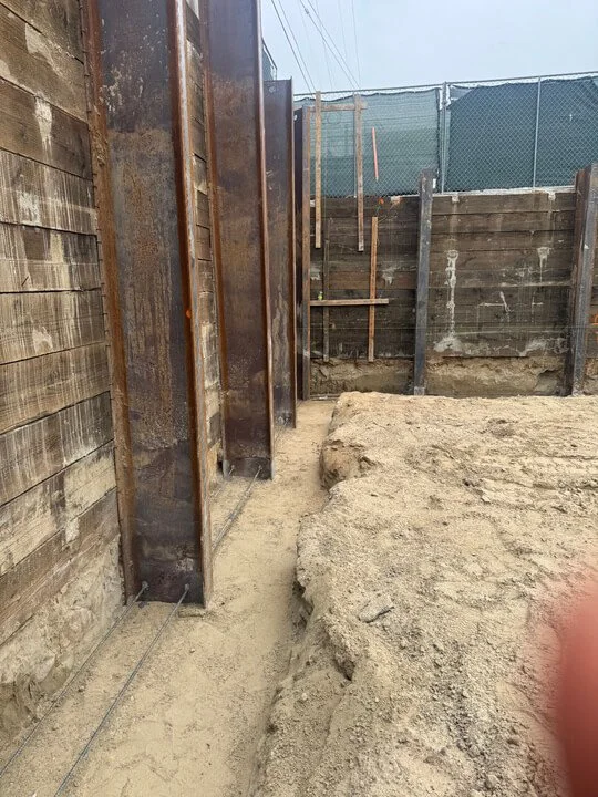 Construction site with steel wall supports and wooden fencing, dirt ground, and construction tools near wooden posts in background.