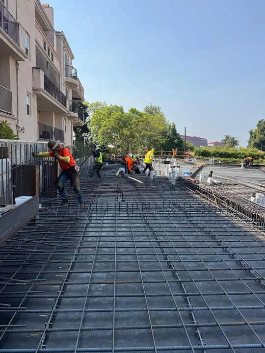 Construction workers installing rebar on a building's concrete floor with in an urban area, a tree, and a clear blue sky in the background.