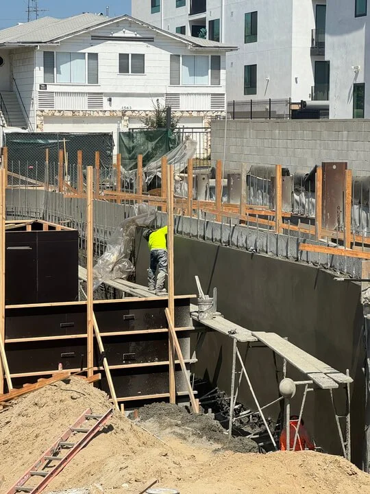Construction worker in a yellow safety vest working on a building foundation, with wooden formwork and concrete structures, in an urban area with houses and apartment buildings nearby.